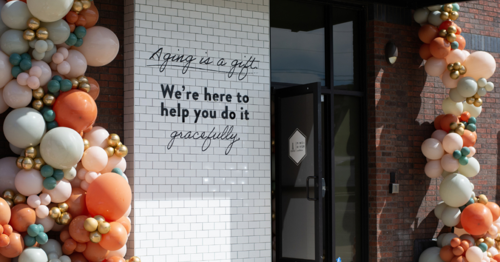 Entrance of Pine & River Aesthetics decorated with elaborate fall balloon garlands in orange, peach, cream, sage green, and gold. A white tiled wall displays the message, “Aging is a gift. We’re here to help you do it gracefully,” beside the glass entrance doors.