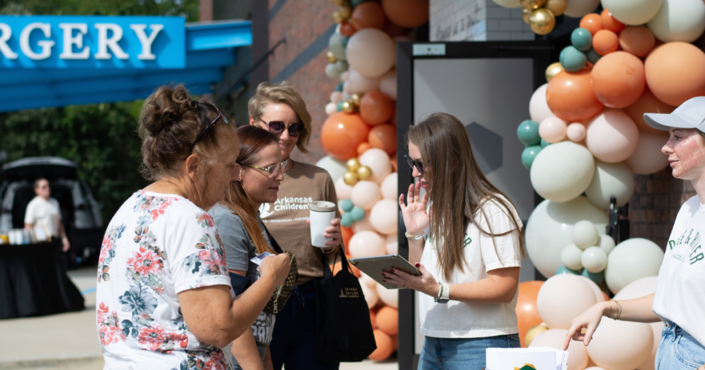 Guests checking in at Pine & River Aesthetics’ 3rd Annual Fall Event. A team member wearing a white Pine & River T-shirt and sunglasses assists attendees with a clipboard outside the entrance, surrounded by decorative balloon garlands in peach, orange, cream, green, and gold.